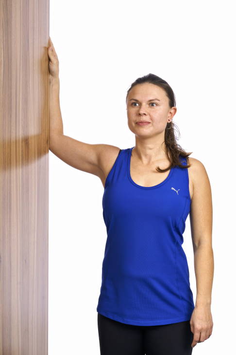 Ausgangsstellung am Türrahmen. © Gelenk-Klinik Frau steht mit angewinkeltem Arm im Türrahmen.
