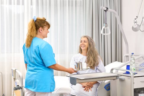 Nach dem Eingriff ermöglicht ERAS eine schmerzfreie und aktive Genesung. © Gelenk-Klinik Mitarbeiterin serviert einer Patientin Getränke.