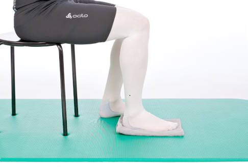 Unter einem Fuß befindet sich ein gefaltetes Handtuch. © Gelenk-Klinik Sitz auf Stuhl, ein Fuß mit Handtuch unterlagert.