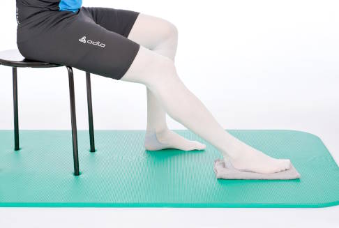 Sitz auf einem Stuhl, ein Fuß schiebt nach vorne. © Gelenk-Klinik Sitz auf einem Stuhl mit Handtuch unter dem rechten Fuß.